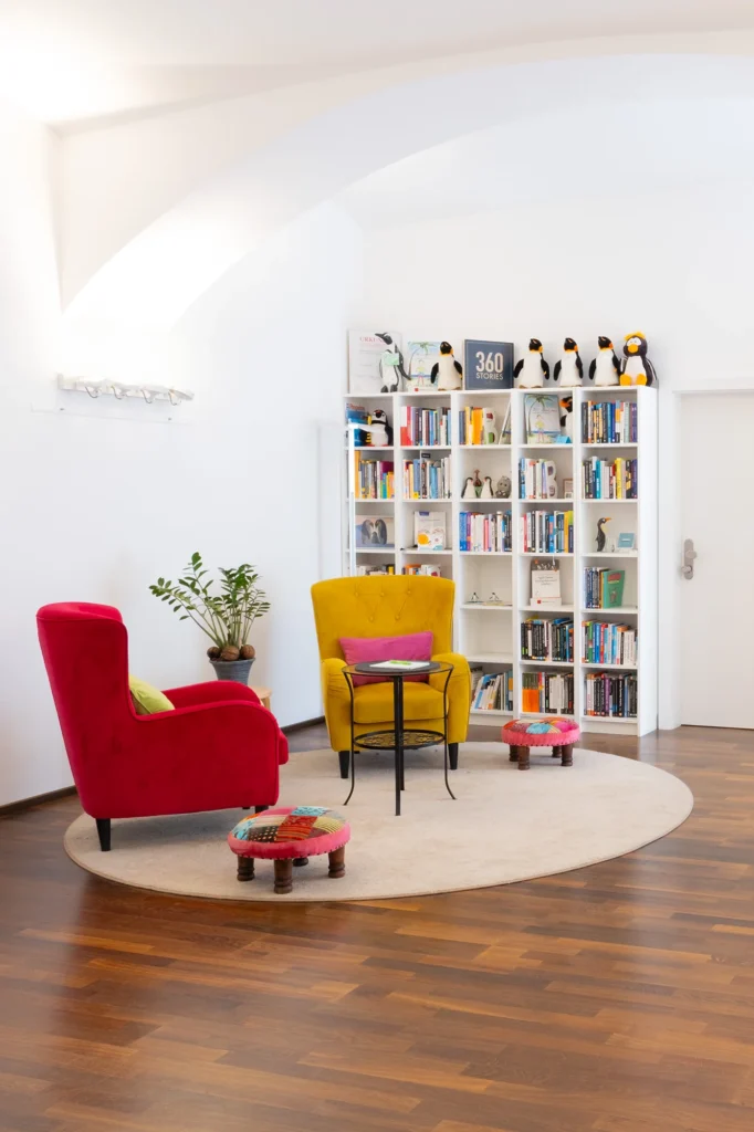 Gemütliche Sitzecke bei SeminarFREUNDE mit rotem und gelbem Sessel, Bücherregal, bunten Hockern und Pinguin-Dekoration